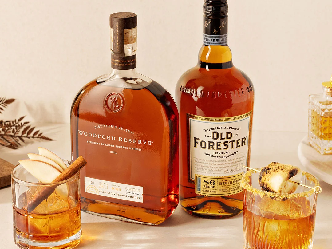 A soft, golden-hued photograph of a bottle of Woodford Reserve Whiskey, and a bottle of Old Forester Whiskey. In front is a Fall Apple Old Fashioned cocktail, and a S'more Old Fashioned cocktail with a roasted marshmallow on a skewer.